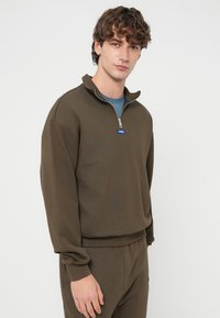 Καφέ φούτερ με φερμουάρ, ψηλό γιακά, ριμπ μανσέτες και λογότυπο. Απαλή υφή, άνετη εφαρμογή, φορεμένο με ταιριαστό παντελόνι.