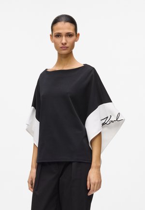 Femme portant un haut ample noir et blanc à manches larges avec "Karl" brodé sur la manche droite, assorti à un pantalon noir.