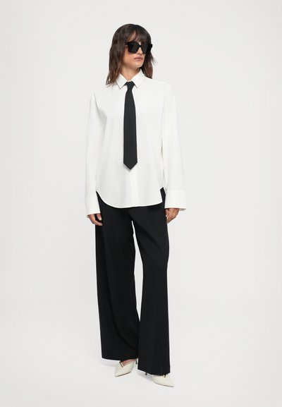 Camicia bianca a maniche lunghe con cravatta nera, abbinata a pantaloni neri a vita alta e gamba larga, e décolleté a punta color crema. Indossati occhiali da sole neri.