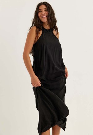 Donna sorridente, indossa un vestito midi nero senza maniche con scollo arricciato, tiene l'orlo del vestito con una mano.