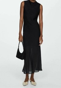 Zwarte mouwloze jurk met een hoge halslijn, vloeiende stof die doorzichtig is aan de zoom, gecombineerd met een zwarte handtas en beige instapschoenen.