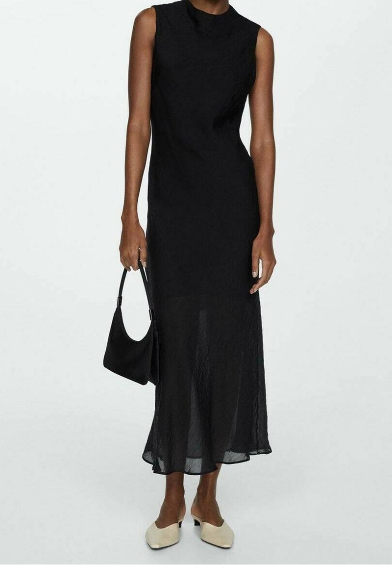 Zwarte mouwloze jurk met een hoge halslijn, vloeiende stof die doorzichtig is aan de zoom, gecombineerd met een zwarte handtas en beige instapschoenen.