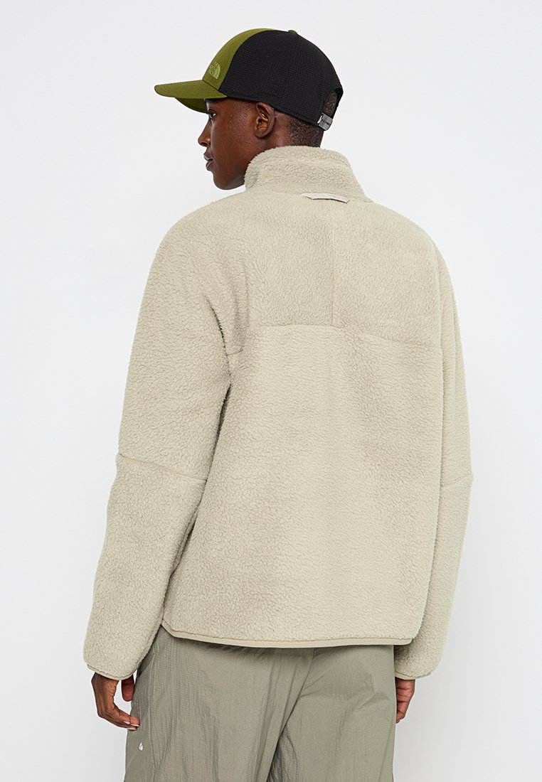 Personne portant une veste en polaire beige, un pantalon vert olive et une casquette de baseball bicolore verte et noire, de dos sur un fond blanc uni.