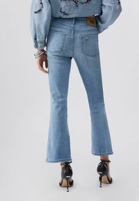 Femme portant un jean évasé bleu clair, des sandales à talons noirs et un haut bleu à manches longues avec des détails en dentelle, vue de dos.