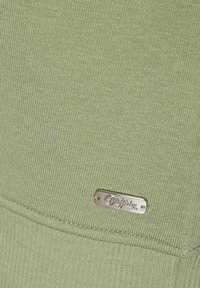 Tela acanalada de color verde claro con una textura sutil. Presenta una etiqueta metálica plateada con el nombre de la marca "Buffalo" cerca del dobladillo.