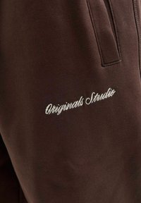 Smeđe dukserice od mekog materijala s diskretnim, bijelim izvezenim logotipom "Originals Studio" sa strane.