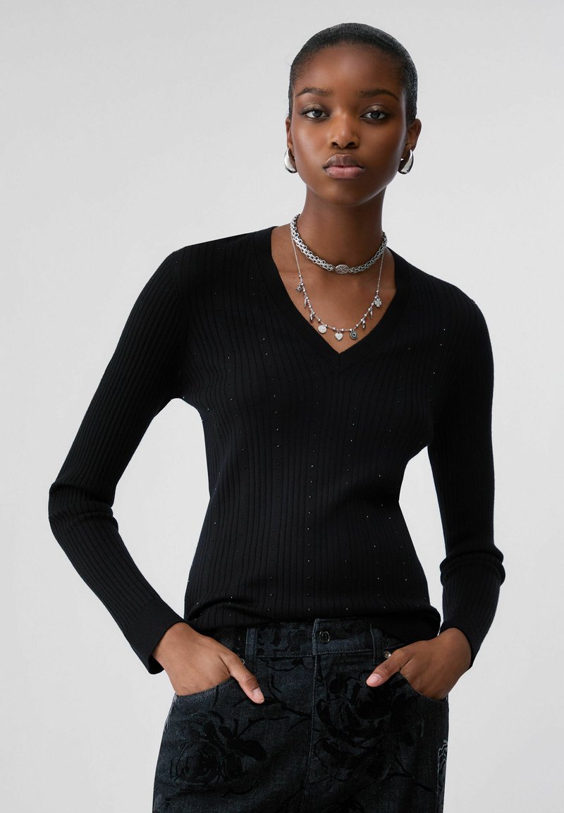 Jeune femme portant un pull noir côtelé à col en V et un pantalon sombre à motifs floraux, des boucles d'oreilles créoles argentées et des colliers argentés superposés.