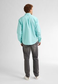 Camicia azzurra chiara a maniche lunghe con polsini a bottoni, dettaglio di piega sulla schiena, indossata con jeans grigi e scarpe bianche. Design semplice e casual.