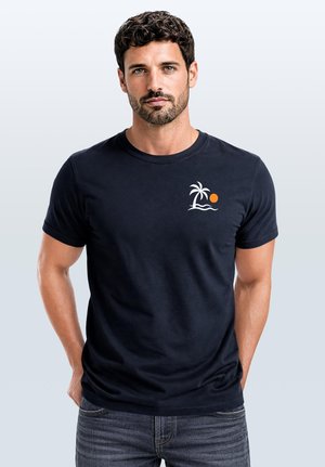 Man with dark curly hair and beard wears black t-shirt with small white palm tree and orange sun graphic, standing against plain background.