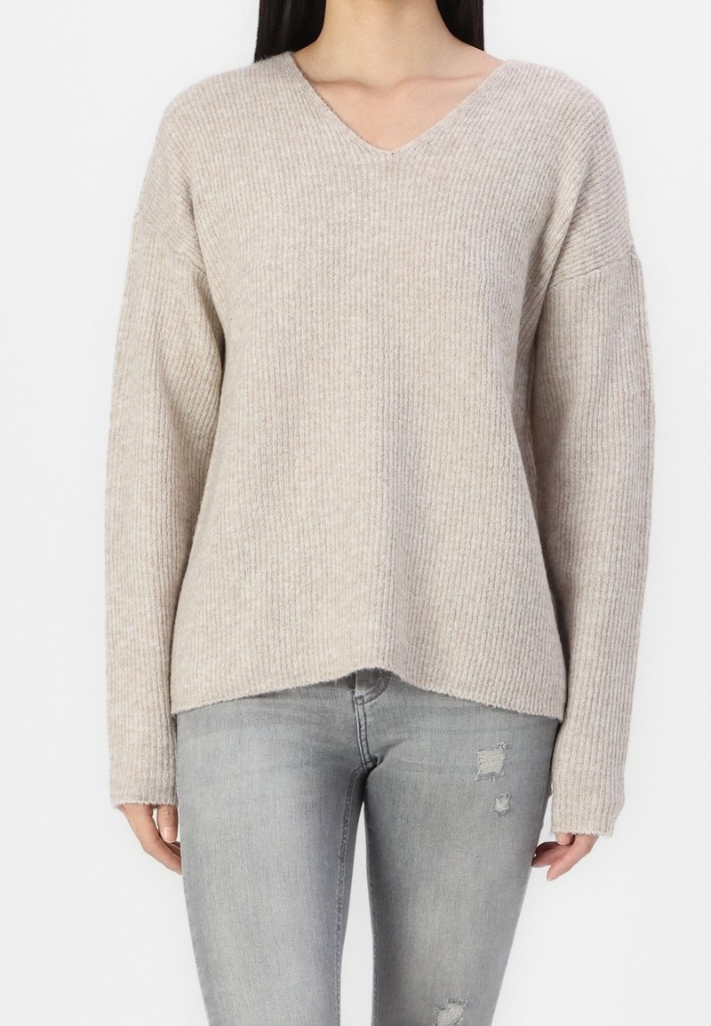 Femme portant un pull beige à col en V côtelé à manches longues et un jean gris clair déchiré, sur un fond blanc.
