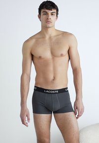 Graue Boxershorts mit einem schwarzen Lacoste-Markenbund. Der Stoff ist glatt und hat ein figurbetontes Design, das flache Nähte aufweist und keine sichtbaren Muster hat.