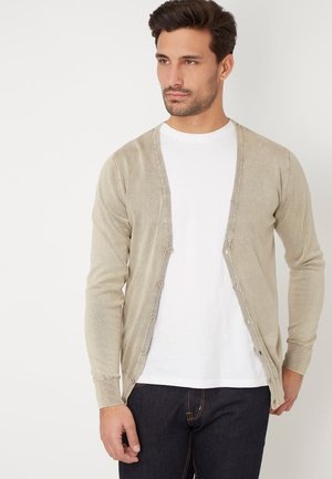 Cárdigan beige con escote en V, cierre de botones y puños acanalados, llevado sobre una camiseta blanca, combinado con jeans oscuros. Textura de tela suave.