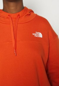 Personne portant un sweat à capuche orange avec des cordons de serrage et un logo blanc The North Face sur le côté gauche de la poitrine.