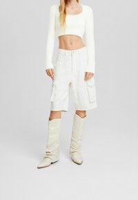 Bílý zkrácený svetr s dlouhými rukávy, spárovaný s bílými cargo šortkami s dvěma bočními kapsami. Outfit doplňují béžové kozačky po kolena.
