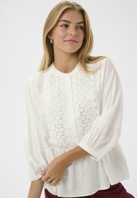 White blouse featuring a button front, patterned embroidery down the center, and puffed long sleeves with cuff detailing. Soft fabric texture.