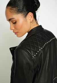 Jauna moteris su tamsiais plaukais, suapvalinta į kuodą, dėvi mažus auksinius žiedus ir juodą pintą odinį švarką su sidabrinėmis smeigėmis ant pečių.