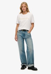 Hvid t-shirt med korte ærmer og "Superdry" logo i guld, parret med lyseblå løse jeans, opsmøget ved anklerne, båret med sorte sko.