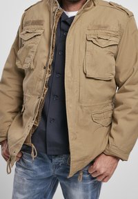 Mężczyzna w odpiętej beżowej kurtce na ciemnej koszuli zapinanej na guziki i wyblakłych niebieskich dżinsach, ręce swobodnie opuszczone wzdłuż ciała na jednolitym tle.