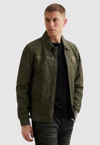 Mladý muž v zelenej koženej bunde so zipsom a detailmi s nášivkami, v čiernom pulóvri a tmavých džínsoch, pozerajúci sa doľava s rukami čiastočne vreckách.