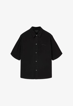Chemise noire à manches courtes avec col, présentant une patte de boutonnage sur le devant et un marquage discret sur le côté gauche de la poitrine. Texture lisse, coupe décontractée.
