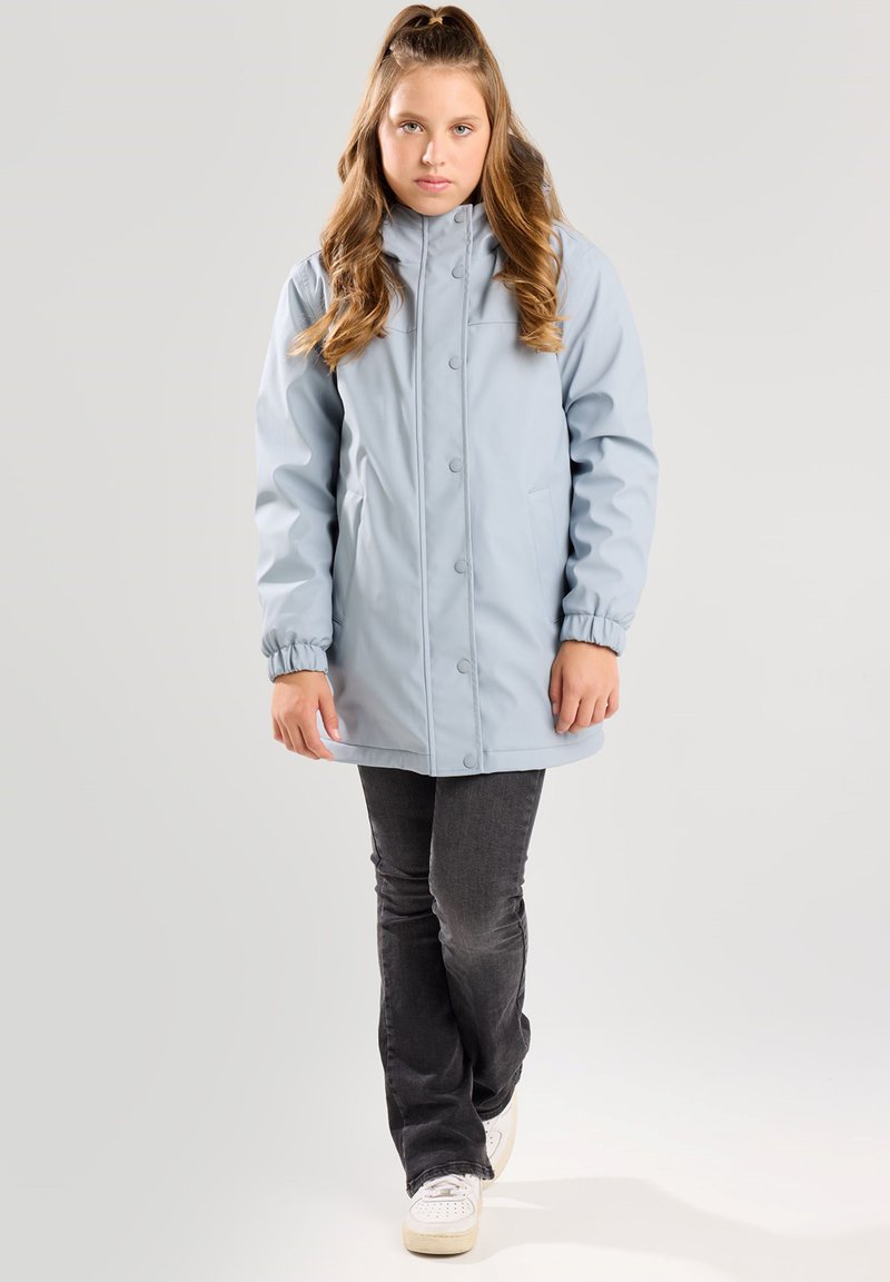 Junges Mädchen mit langen Haaren trägt eine hellblaue geknöpfte Jacke, dunkelgraue Jeans und weiße Turnschuhe, stehend vor einem einfarbigen Hintergrund.