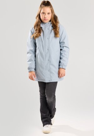 Parka impermeable de color azul claro con cuello alto, botones a presión y puños elásticos, combinado con jeans oscuros y zapatillas blancas.