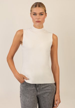 Weißes ärmelloses Oberteil mit hohem Kragen, aus geripptem Material bestehend. Kombiniert mit grauen Jeans mit dekorativen Perlenakzenten.