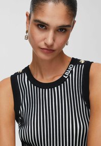 Haut sans manches à rayures noires et blanches avec une texture côtelée, comportant des accents de boutons dorés sur les épaules et un détail de logo.