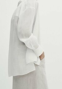 Witte linnen blouse met lange mouwen, een relaxte pasvorm en een drop schouderontwerp. Naaddetail aan de achterkant, gecombineerd met een lichtgrijze rok.