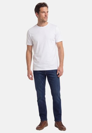 Mann trägt ein einfaches weißes T-Shirt mit kurzen Ärmeln, dunkelblaue Jeans und braune Turnschuhe, steht und schaut zur Seite.