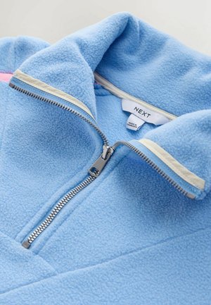 Vaaleansininen fleece-takki, jossa on korkea kaulus, etuvetoketju ja kontrastivärinen kermaisen vaalea reunus vetoketjun ympärillä. Sisäpuolella näkyvä merkki.