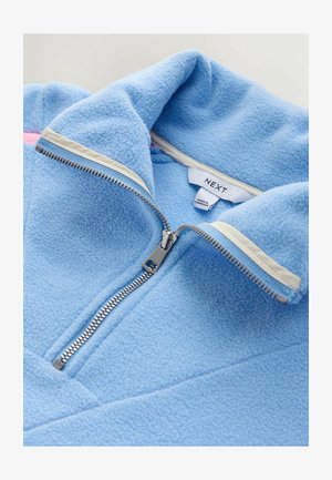 Vaaleansininen fleece-takki, jossa on korkea kaulus, etuvetoketju ja kontrastivärinen kermaisen vaalea reunus vetoketjun ympärillä. Sisäpuolella näkyvä merkki.