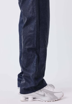 Jeans in denim blu scuro con una vestibilità rilassata, caratterizzati da una texture liscia. Abbinati a scarpe da ginnastica bianche con dettagli argentati.