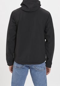 Osoba w czarnej kurtce z kapturem i jasnych niebieskich dżinsach, pokazana od tyłu na jednolitym białym tle.