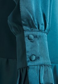 Blusa teal con maniche arricciate e due bottoni rivestiti in tessuto coordinato al polsino. Il materiale appare liscio con una leggera brillantezza.