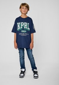 T-shirt oversize bleu marine avec texte imprimé blanc et vert, associé à un jean bleu ajusté et des baskets noires avec accents blancs.