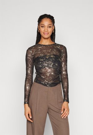 Top negro transparente de manga larga con brillo dorado, que presenta detalles fruncidos y un escote redondo, combinado con pantalones marrones de cintura alta.
