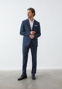 Marineblauwe pak gemaakt van gestructureerde stof, met een getailleerde pasvorm, notch-lapels, twee voorkruiden en een wit overhemd eronder.