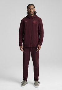 Tuta sportiva bordeaux con giacca con cappuccio zip e pantaloni abbinati. Presenta un logo ricamato e cuciture di accentuazione rosa. Sneaker grigie.