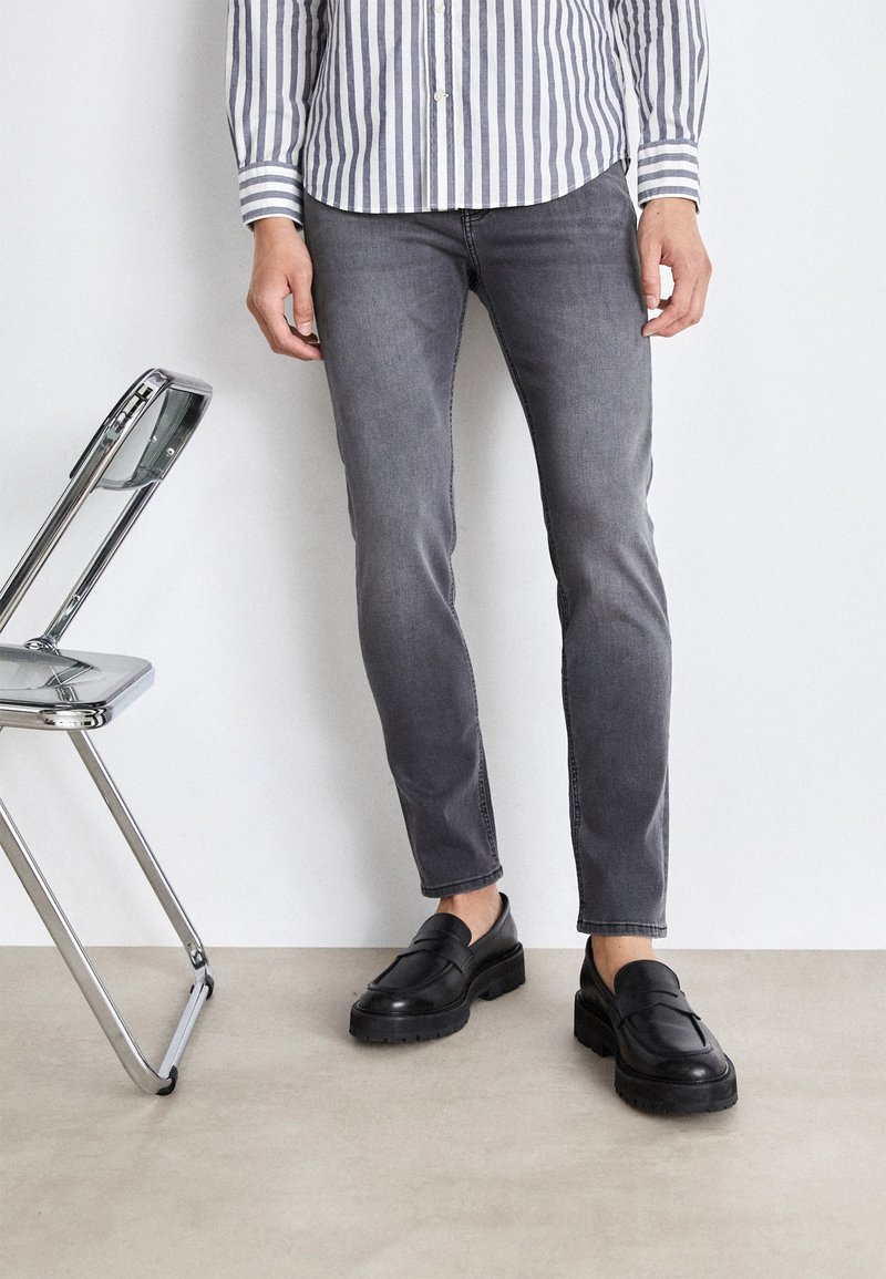 Homme portant un jean gris slim, des mocassins noirs et une chemise à rayures à boutons, debout à côté d'une chaise pliante en chrome et transparent.