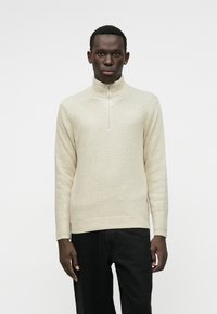 Pullover lavorato a maglia crema con colletto alto, dotato di una cerniera a un quarto e un motivo testurizzato. Abbinato a pantaloni neri, centrato su uno sfondo chiaro.