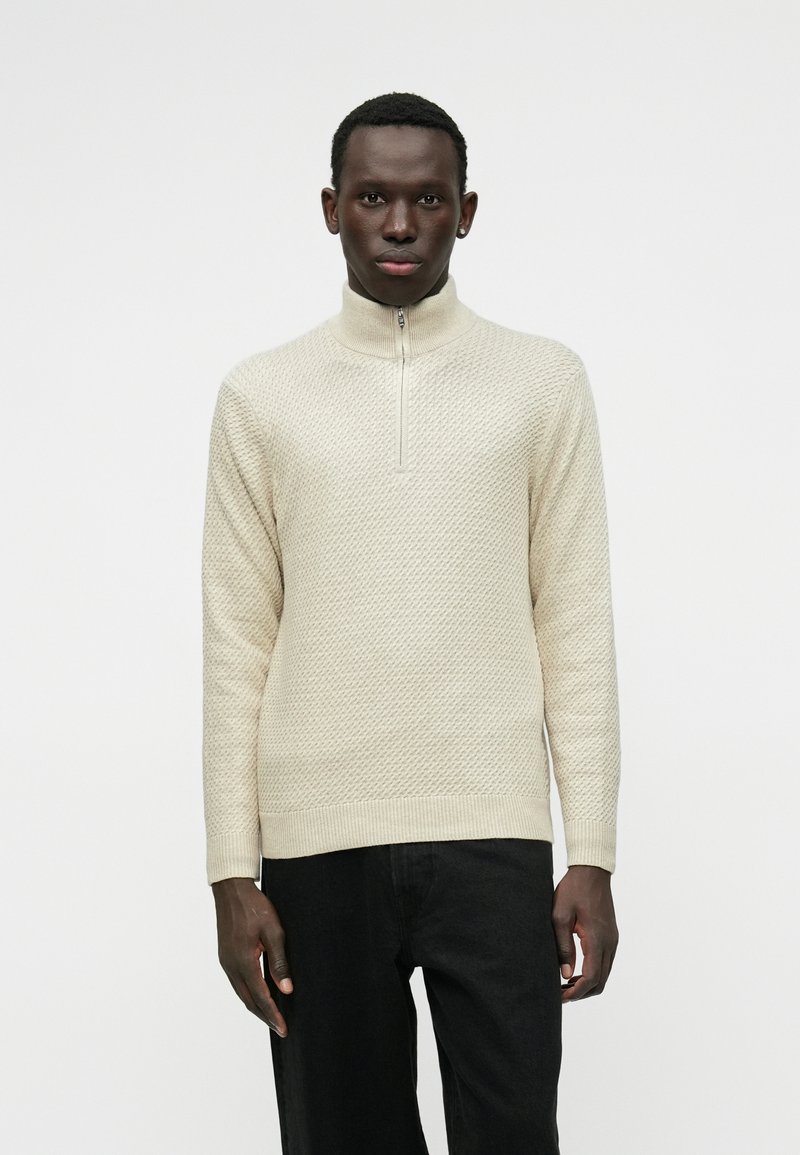 Pullover lavorato a maglia crema con colletto alto, dotato di una cerniera a un quarto e un motivo testurizzato. Abbinato a pantaloni neri, centrato su uno sfondo chiaro.