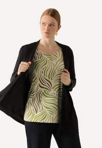 Černý cardigan s otevřeným předním dílem přes krémový top s zelenými vlnitými listovými vzory. Modelka má na sobě tmavé kalhoty a zlaté geometrické náušnice.