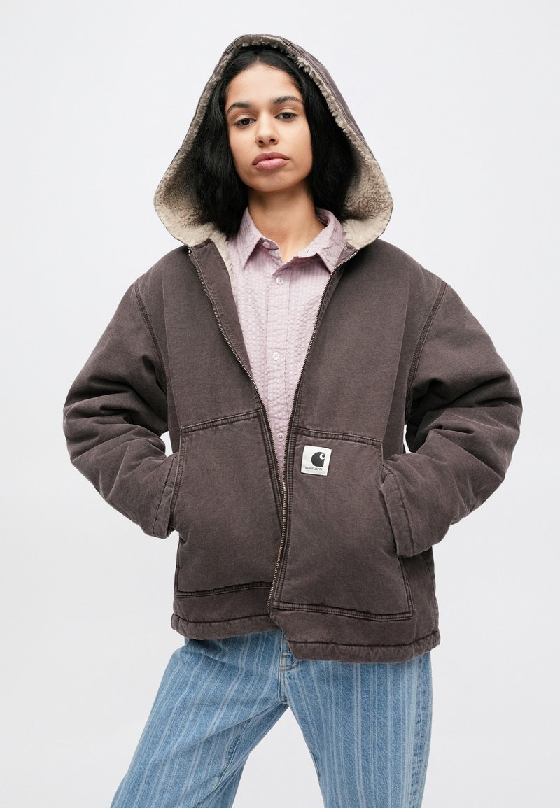 Dark brown hooded jacket with a soft inner lining, front pockets, and logo patch. Worn over a light pink button-up shirt and blue striped jeans.