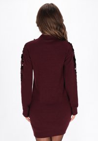 Donna con lunghi capelli castani che indossa un vestito maglione attillato di un rosso scuro con paillettes nere sulle maniche, in piedi di spalle su uno sfondo bianco.