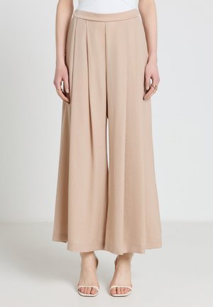 Beige Hosen mit weitem Bein und vorderen Falten, getragen mit weißen Sandaletten mit Absatz, gezeigt von der Taille bis zu den Füßen vor einem einfarbigen Hintergrund.