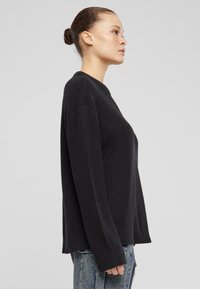Schwarzer Strickpullover mit einem strukturierten Rippmuster, langen Ärmeln und rundem Halsausschnitt. Der Saum ist hinten etwas länger.