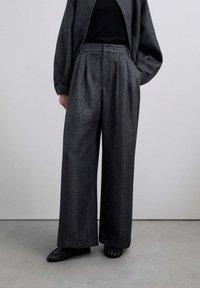 Pantalones de pierna ancha en tela gris oscuro, con pliegues y cintura alta. Combinados con un top negro y zapatos planos negros.