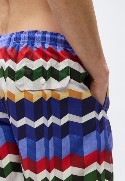 Κοντινό πλάνο ενός ατόμου που φοράει πολύχρωμα σορτς με γεωμετρικό μοτίβο, με τσέπη με καπάκι και το χέρι στην πλαϊνή τσέπη.