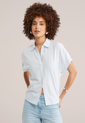 Naine lokkis juustega, kandes hõbedaid rõngakujulisi kõrvarõngaid, hele sinist lühikeste varrukatega nööpidega särk, helesiniseid teksaseid ja hõbedast käevõru.